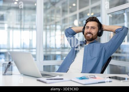 Smiling man enjoying a moment of relaxation while listening to music with headphones, leaning back in his office chair during a successful work break at a modern coworking space Stock Photo