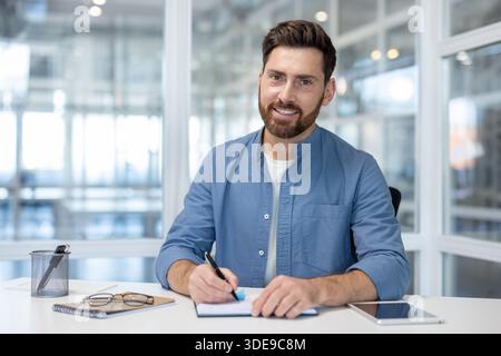 Confident bearded professional in a blue shirt smiles at camera while writing notes at his desk in a bright modern office, focused and productive on paperwork and tablet planning Stock Photo