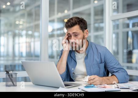 Overworked businessman sitting at an office desk, holding his head and a tissue, feeling exhausted and having a headache from stress and fatigue during a long workday Stock Photo