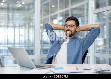 Young man wearing eyeglasses feeling relaxed and smiling, leaning back in his chair with hands behind head, taking a break from working with laptop at modern office Stock Photo
