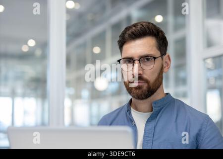 Confident man in glasses and beard looking at the camera, posing in front of a modern office background, reflecting professionalism, focus, and corporate success while working Stock Photo