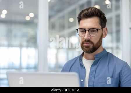 Young man with beard and glasses concentrating on his work, using a laptop in a bright contemporary workplace, representing dedication and professional activity Stock Photo