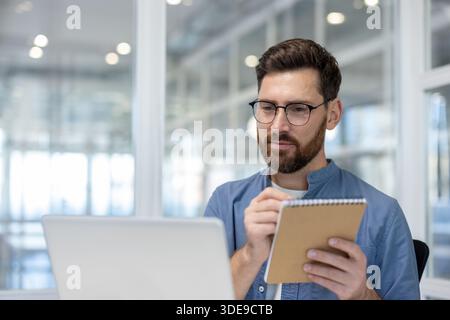 Entrepreneur with glasses concentrating on data while taking notes in a spiral notebook, attending to business tasks in a bright, modern office with a laptop Stock Photo