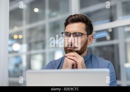 Professional bearded man in glasses sitting at his laptop in a bright contemporary office, reflecting deeply with clasped hands, focusing on a complex business challenge or problem-solving Stock Photo