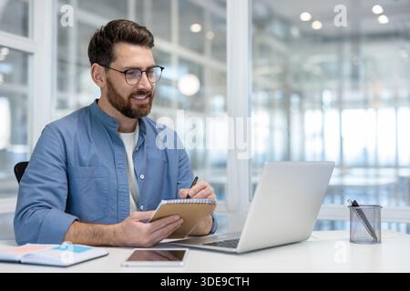 Positive bearded businessman working on a laptop, smiling and writing in a notebook while participating in a virtual conference or remote work session in a modern office Stock Photo
