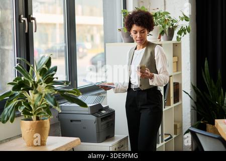 A young African American manager reviews documents while checking her phone in a stylish office. Stock Photo