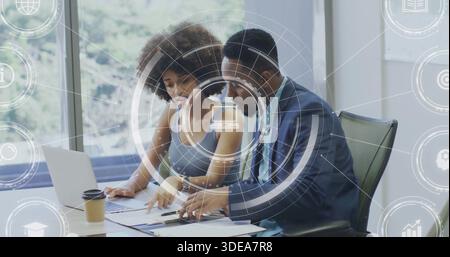 Collaborating woman in sleeveless top and male in suit reviewing papers in office laptop coffee HUD Stock Photo