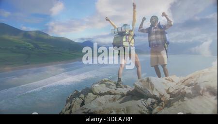Standing hikers raising arms on cliff overlooking sandy bay, in shorts and packs with pad Stock Photo