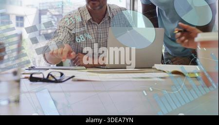 Discussing three colleagues in business wear collaborating at table in office, with laptop, charts Stock Photo