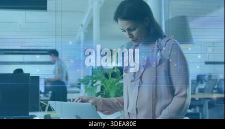 Standing woman in pink blouse typing on laptop at open office with plant and blue overlay Stock Photo