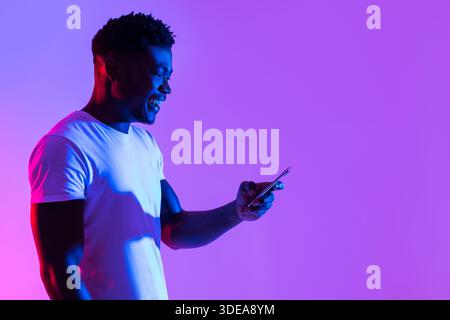 Joyful Young Black Man Uses Smartphone in Vibrant Neon Light While Working or Studying Stock Photo
