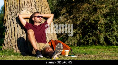 Urban man relaxing in city park, sitting near tree with hands behind head. Stock Photo