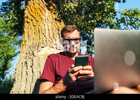 Low view of man using smartphone and laptop sitting near tree outdoors Stock Photo