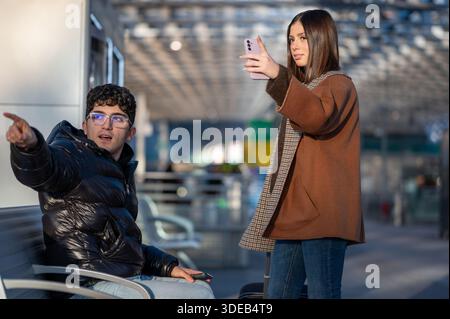 Young couple traveling in a modern train station, the man pointing while sitting and the woman standing holding a mobile phone looking for information Stock Photo