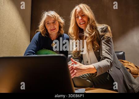 Two engaged women collaborate over a laptop in a warm, inviting office setting. They share smiles and insights, creating a sense of connection and tea Stock Photo