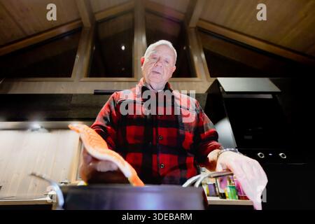 Elderly man in a red plaid shirt is cooking salmon in a contemporary kitchen, surrounded by wooden elements and bright lighting, highlighting culinary Stock Photo