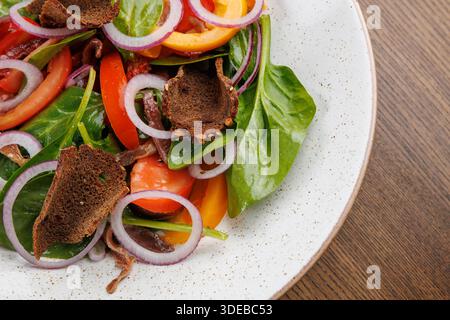 A colorful salad features spinach, ripe tomatoes, sliced peppers, red onions, and pieces of dark bread. It is served on a white plate set on a wooden Stock Photo