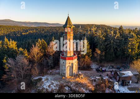 Stepanka lookout tower stands tall in the Jizera Mountains during winter. Visitors gather at the base as the sun sets over the landscape, creating a warm glow in the sky. Stock Photo