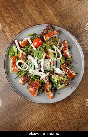 A fresh salad is served with grilled chicken, mixed greens, cherry tomatoes, and white noodles. The dish is presented on a gray plate, set against a w Stock Photo