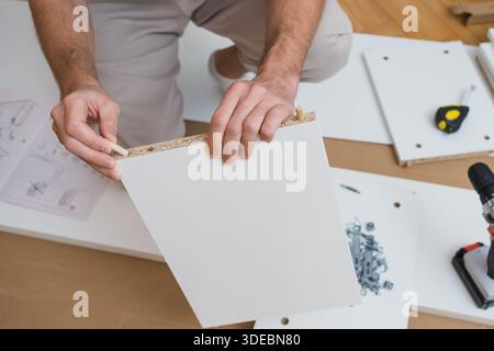 man carefully aligns two furniture panels with dowels on a wooden floor surrounded by tools, hardware, and assembly instructions in a bright room Stock Photo