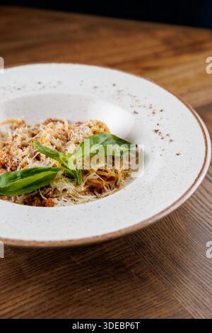 A serving of spaghetti topped with meat sauce, grated cheese, and fresh basil leaves sits on a round white plate. The wooden table enhances the dining Stock Photo