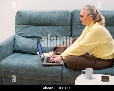 A relaxed senior woman is engaged in work on her laptop, sitting cross-legged on a soft, blue couch. A warm cup and a small notebook are nearby, creat Stock Photo