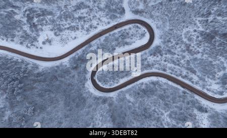 Vehicles traveling through dense, snow-covered forest and a winding road seen from above. Stock image with a winter travel and transportation theme. Stock Photo