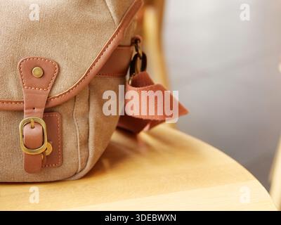 Close up of vintage canvas messenger bag with leather straps and brass buckle on wooden chair, retro shoulder bag detail, stylish travel accessory. Stock Photo