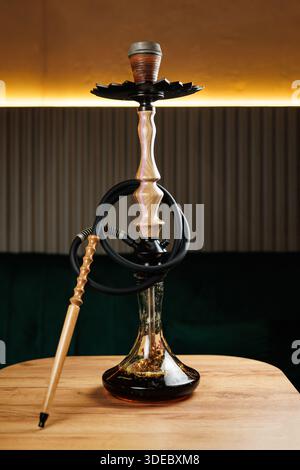 A traditional hookah sits on a wooden table, surrounded by soft lighting in a lounge. Smoke curls gently from the top, creating an inviting atmosphere Stock Photo