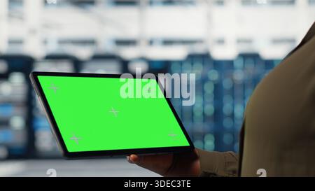 Server room admin uses isolated screen tablet to increase supercomputers workload capacity. Data center worker configures gear with chroma key device notebook, improving processing speed, camera B Stock Photo