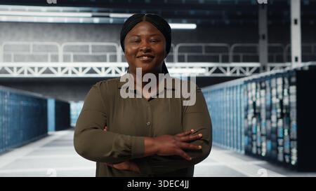 Portrait of smiling data center admin conducting security audit to protect against cyber threats. Happy supervisor walking in server room, pleased by compliance with industry standards, camera B Stock Photo