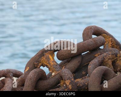 Close up of large corroded iron chains with orange rust texture piled against a blurred blue sea background at a harbor Stock Photo
