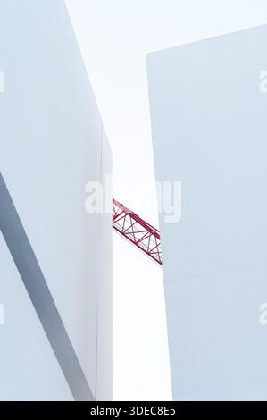 Minimalist white modern architecture with geometric walls and red construction crane visible between structures, abstract urban composition. Stock Photo