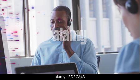Speaking seated man wearing light blue shirt using headset microphone in modern office, with laptop Stock Photo