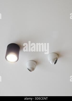 Modern interior ceiling with illuminated black spotlight and white security camera dome sensors installed on plain surface offering safe feeling in mi Stock Photo