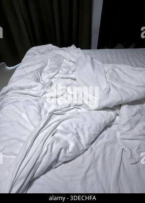 Close up view showing unmade white bed with wrinkled duvet blanket and messy sheet reflecting lazy morning atmosphere inside cozy bedroom Stock Photo