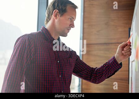 Whiteboard, sticky notes and businessman in office with research for creative project at startup. Reading, paper and male magazine editor with Stock Photo