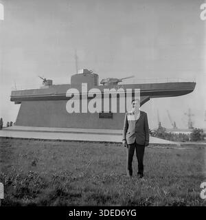 Izmail, Odessa region, Ukrainian SSR, Soviet Union, 1975. A male tourist posing for a souvenir photo in front of the famous Monument to the Armored Motor Gunboat BK-134 near the Danube Commercial Port. The black and white archive image shows a man in a suit standing on the grass, with industrial cranes visible in the distance. The memorial is dedicated to the Danube Flotilla sailors who liberated the city. The shot captures the atmosphere of patriotic tourism and peaceful leisure in the southern city during the stable USSR era. Stock Photo