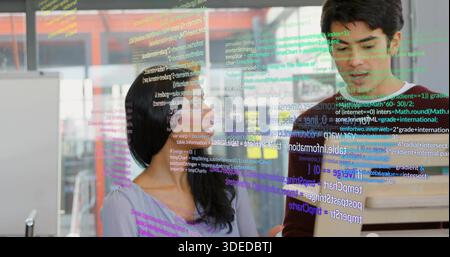 Debugging purple-top woman and green-knit man reviewing code at office with laptop, glass partition Stock Photo