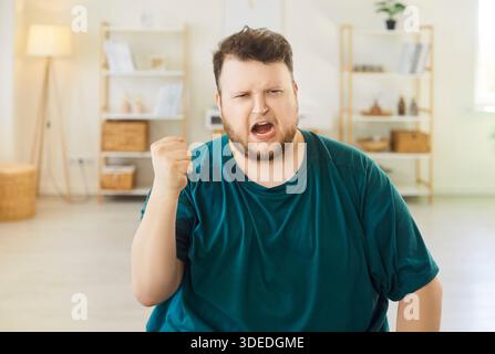 Frustrated man screaming with raised hands at home, expressing stress and rage Stock Photo