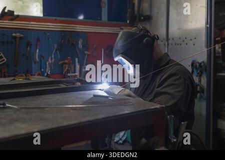 A professional welder is working with metal in an industrial workshop surrounded by tools and equipment. Stock Photo