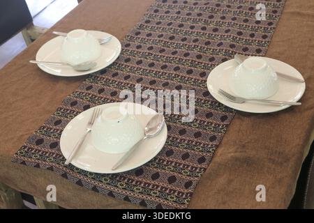 View of white ceramic dish set with bowl and stainless cutlery on patterned fabric runner at wooden dining table creating welcoming and organized atmo Stock Photo
