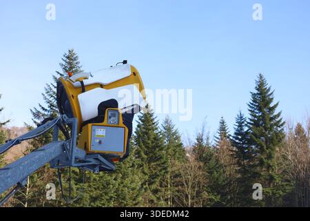 Side view of a white and yellow snow cannon with forest and blue sky background Stock Photo