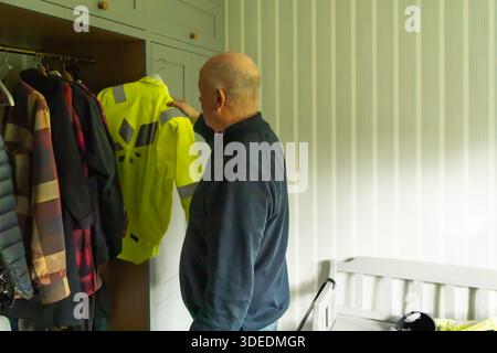 Older male is inspecting a high-visibility jacket in a tidy closet filled with diverse garments, reflecting readiness for outdoor activities and safet Stock Photo