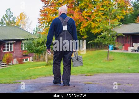 Elderly man in dark overalls walks away from charming homes, holding toolbox, with colorful autumn trees creating a serene atmosphere in the backgroun Stock Photo