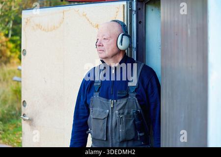 Elderly male worker in dark overalls stands outside workshop, wearing protective headphones, observing environment with a focused demeanor, reflecting Stock Photo