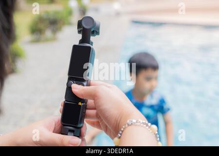 Young woman holding a compact camera, action camera to taking photos and videos of her family playing in the swiming pool. Selective focus. Stock Photo