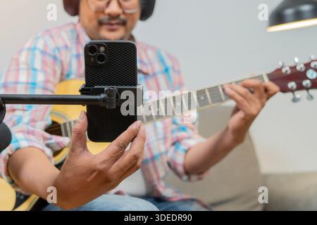 Phone, guitar tutorial and man with a smile online to coach during live streaming lesson. Asian male influencer happy on home sofa, teaching music on Stock Photo