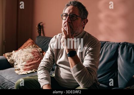 Thoughtful middle aged man sitting on a sofa at home, touching his lips while thinking. Concept of reflection, mental health, stress, decision making Stock Photo