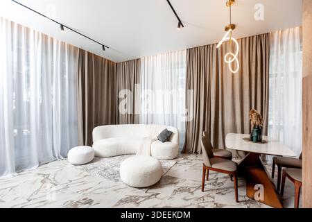A living room with a white couch, a coffee table, and a dining table. The room is decorated with white curtains and a gold lamp. The atmosphere is coz Stock Photo